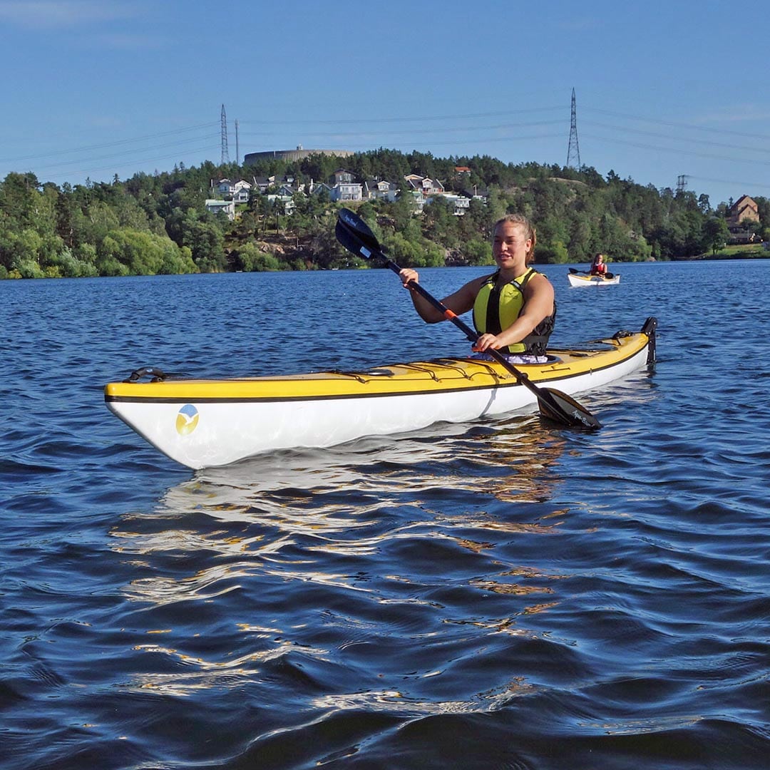 En paddlare i en gul Seabird Expedition-glasfiberkajak ses från sidan på lugnt vatten med skog i bakgrunden.