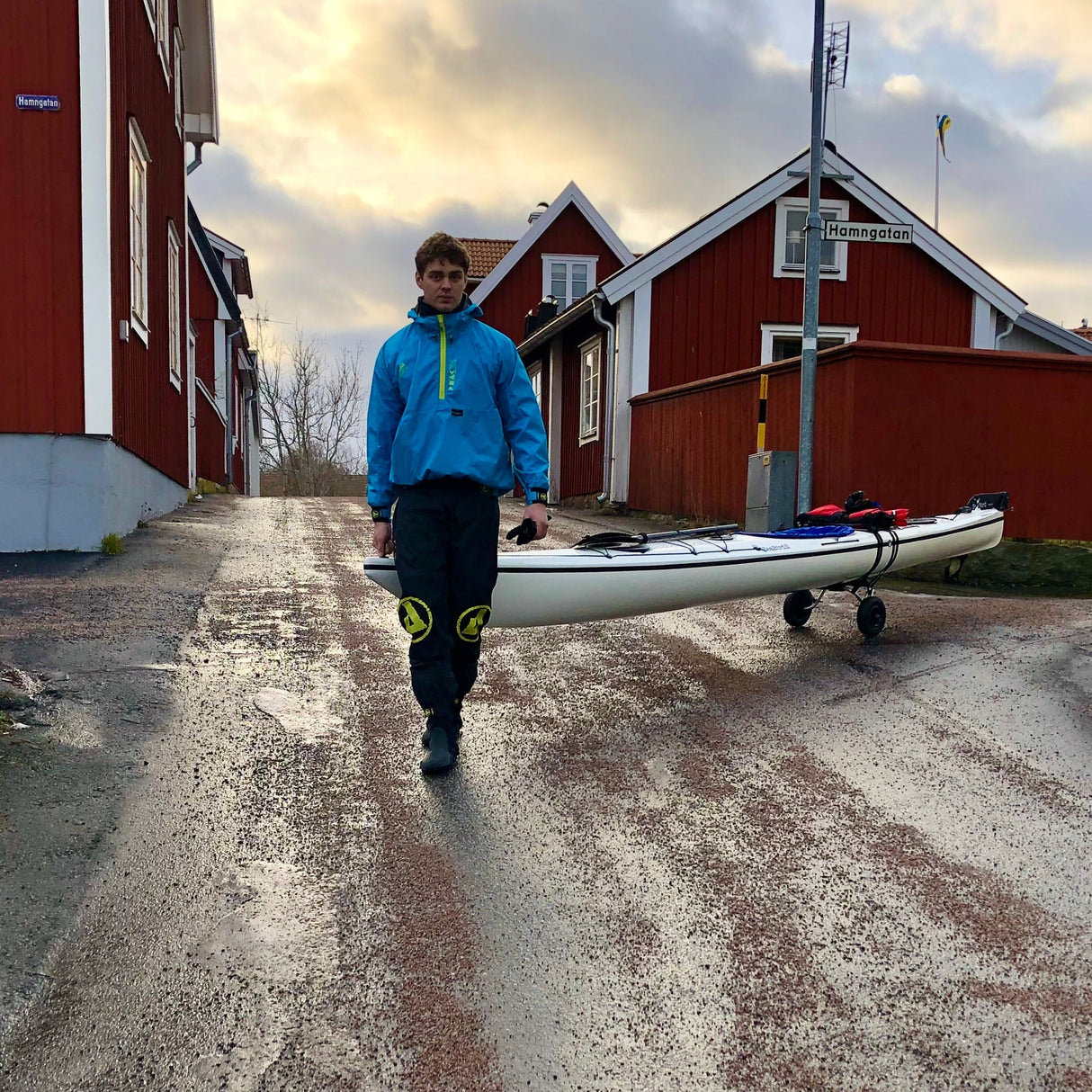 Person i blå paddeljacka drar en vit kajak på en kajakvagn längs en väg med röda hus i bakgrunden