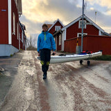 Person i blå paddeljacka drar en vit kajak på en kajakvagn längs en väg med röda hus i bakgrunden